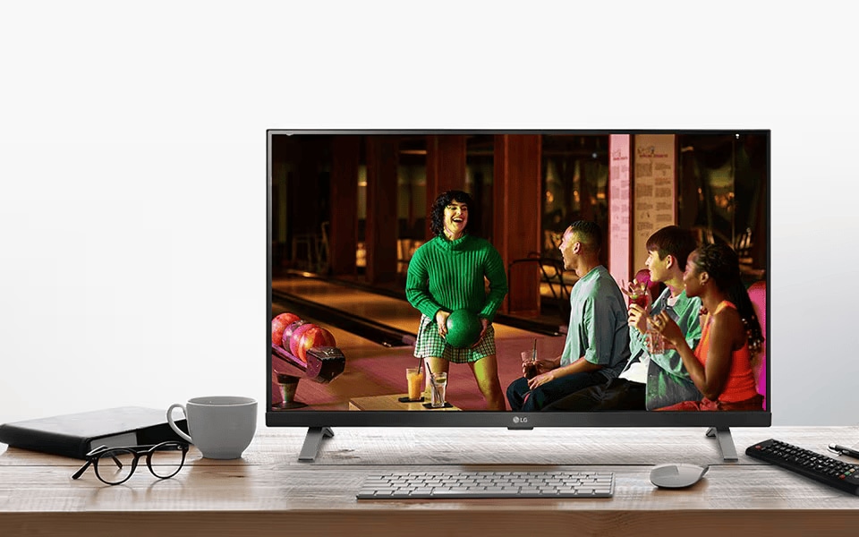 LG Smart TV on a desk displaying vibrant bowling scene, surrounded by office items like keyboard, mug, and glasses.