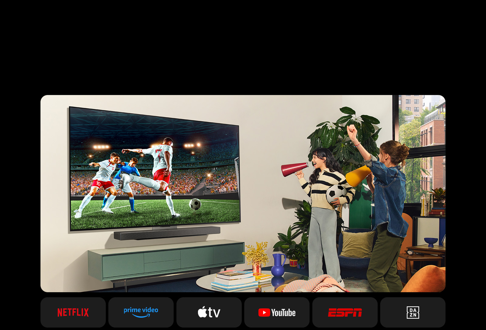 Deux femmes regardent un match de football dans un espace de vie confortable. Une femme tient un ballon de football et les deux soutiennent leur équipe avec des haut-parleurs jaune et rouge. Les logos suivants sont présentés ci-dessous. Netflix, Amazon Prime Video, Apple TV, Youtube, ESPN et DAZN.