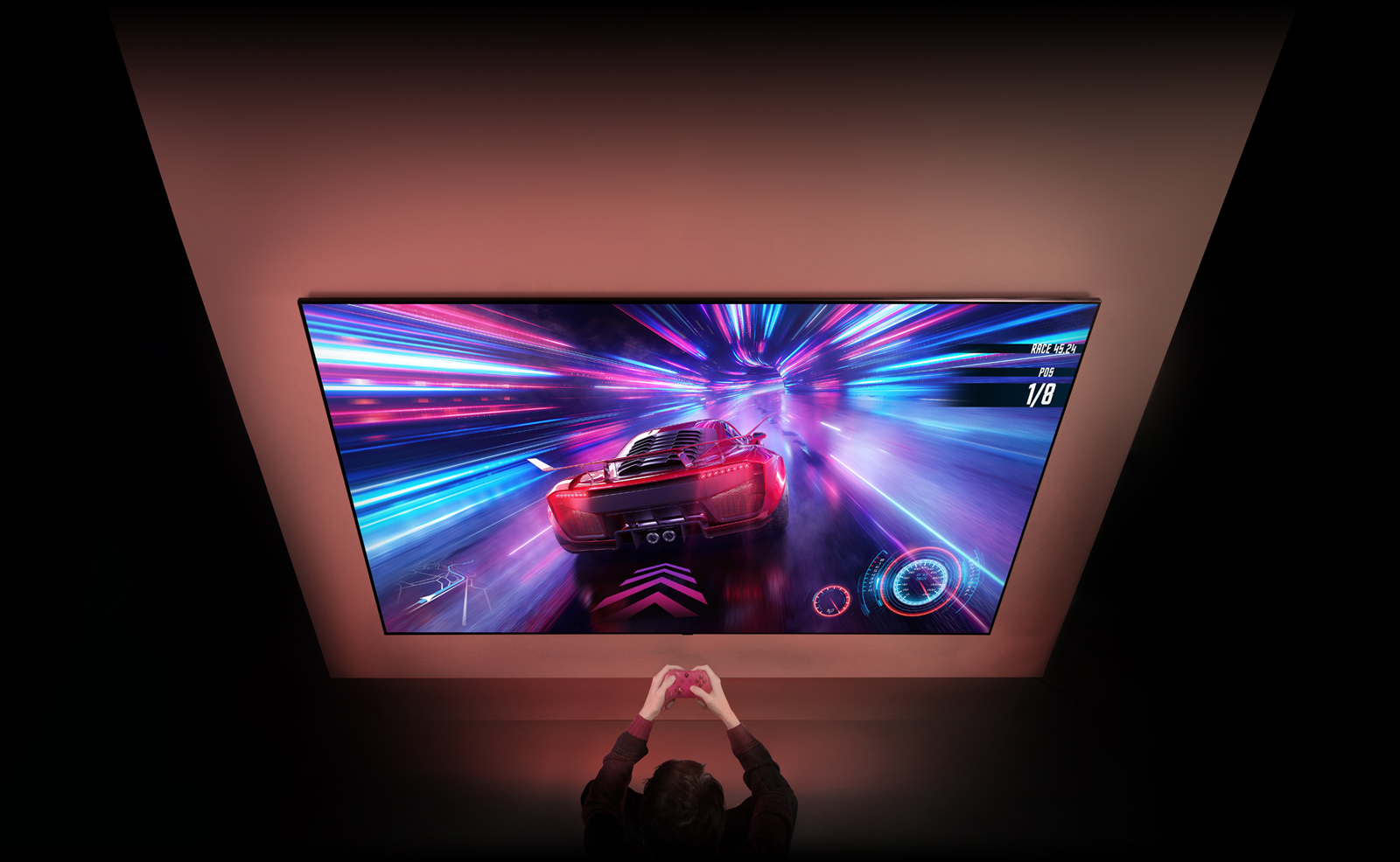 Il y a un grand téléviseur sur le mur et on peut voir un jeu de course de voitures sur l’écran. Devant le téléviseur, on aper?oit les mains et la manette d’une personne concentrée sur son jeu.
