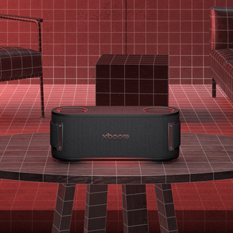 Enceinte LG XBOOM placée sur une table dans une pièce aux tons rouges avec des murs à motifs quadrillés et des meubles modernes