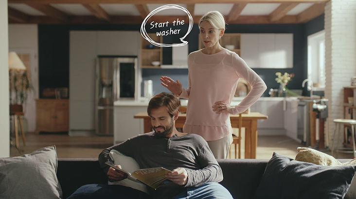 Dans une cuisine intelligente moderne, une femme utilise une commande vocale pour démarrer la machine à laver avec LG ThinQ AI, tandis qu'un homme lit sur le canapé en arrière-plan.