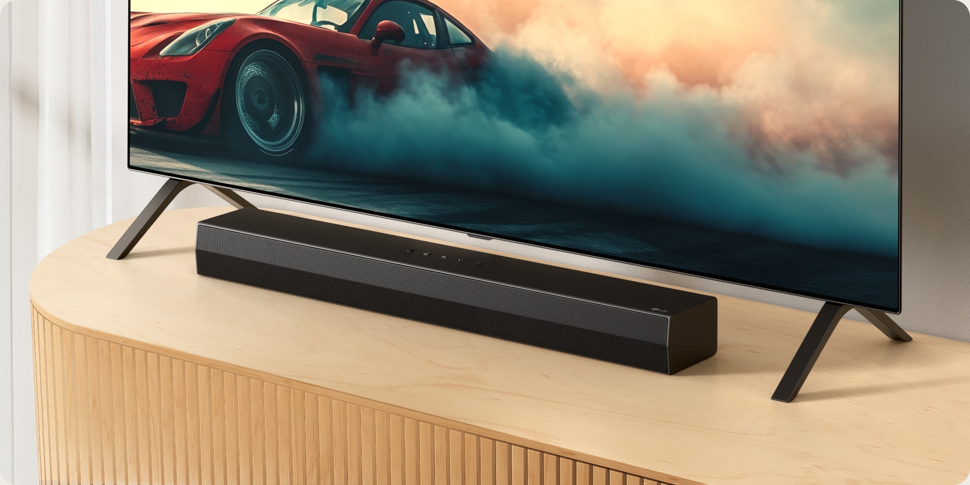 LG Soundbar S20A е поставен на дървена телевизионна конзола, под телевизора и между стойките му. По телевизора се вижда червена кола с пушеци.