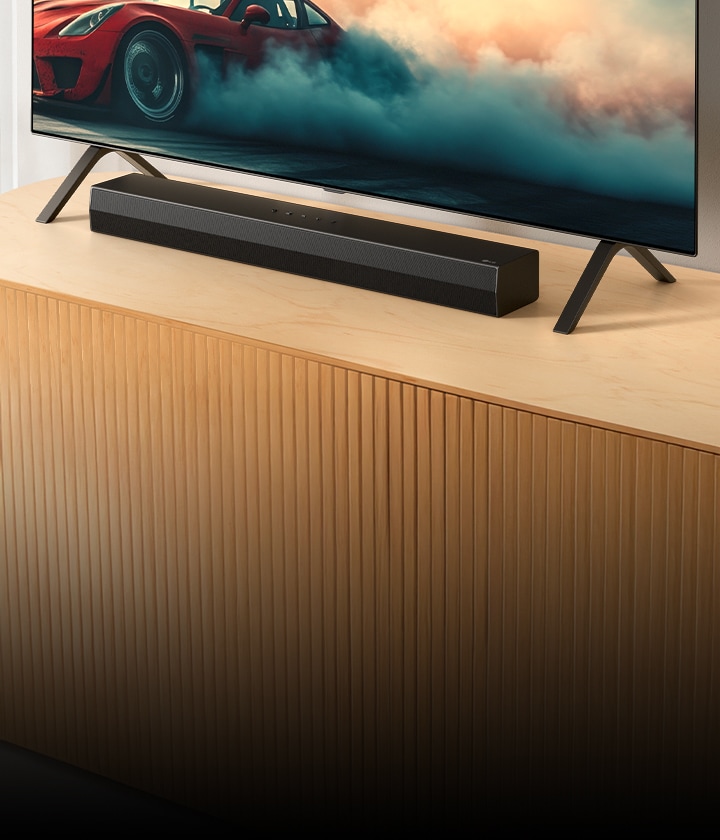 LG Soundbar S20A е поставен на дървена телевизионна конзола, под телевизора и между стойките му. По телевизора се вижда червена кола с пушеци.
