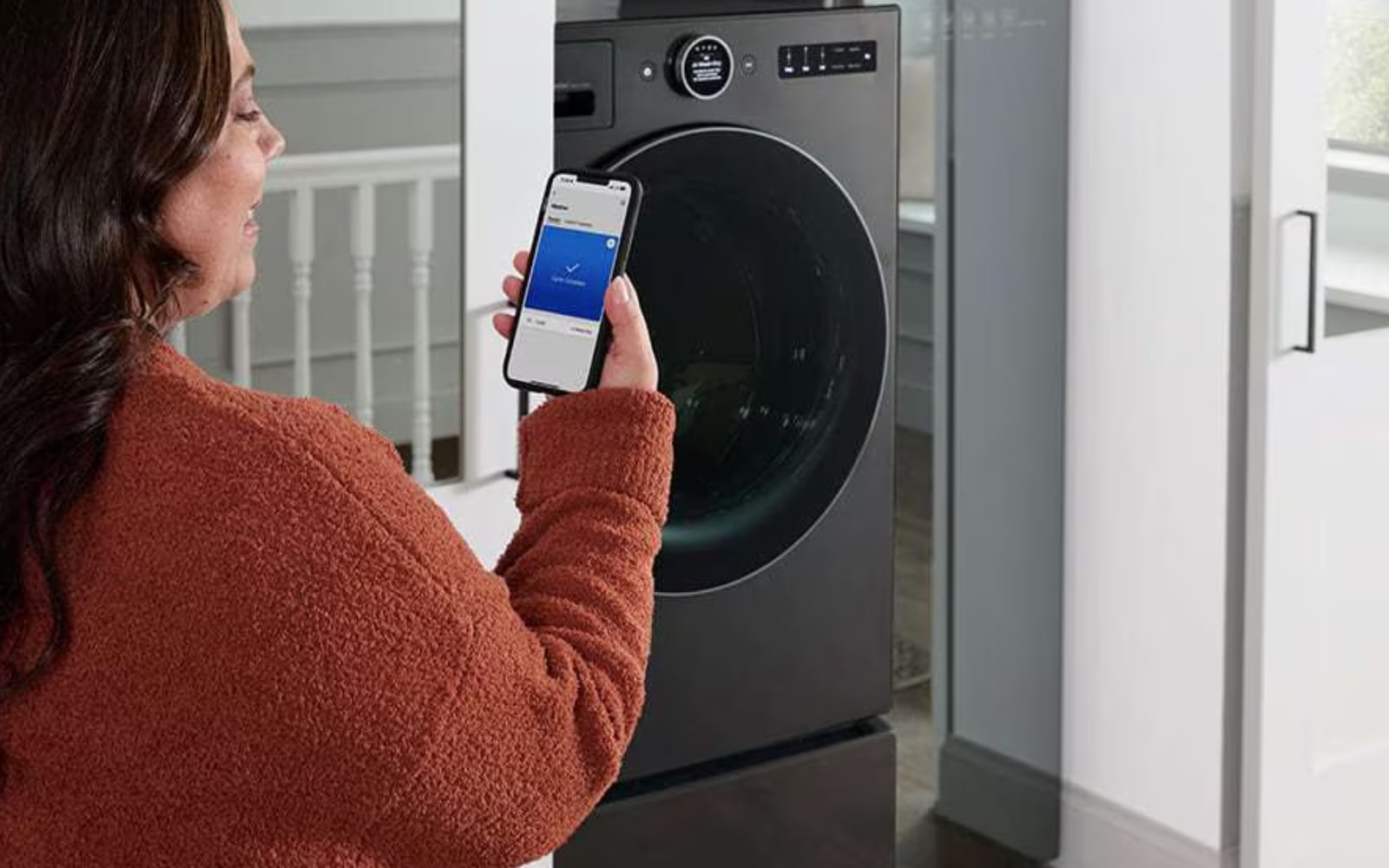 Person using the LG ThinQ? app on their smartphone to control or monitor an LG washing machine. The person appears to be selecting a washing program remotely, highlighting the smart connectivity and convenience features of the appliance.