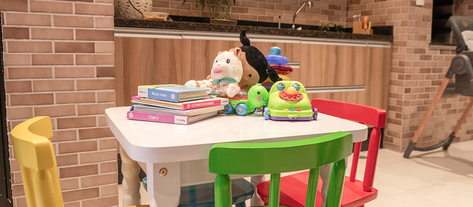 Uma imagem de mesa branca e cadeiras coloridas com amarelo, verde, vermelho. Na mesa est?o bonecas, livros e alguns brinquedos para as crian?as.
