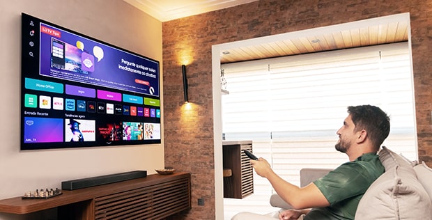 O editor especial Enzo está sentado em um sofá, segurando um controle remoto para operar a TV LG OLED. A fun??o WebOS está sendo exibida na tela.