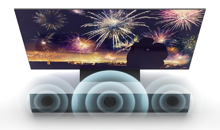 Na vis?o aérea, a TV LG está apoiada no ch?o, e a Sound Bar LG está logo abaixo, num espa?o infinito. Na tela da TV, a silhueta de um casal sobre um fundo com fogos de artifício. Gráficos de ondas sonoras emanam dos alto-falantes central, esquerdo e direito no topo da sound bar.