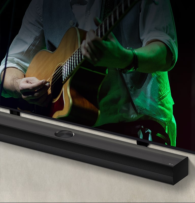 LG Soundbar contra um fundo preto revela seu design come?ando no canto esquerdo e depois se expande para mostrar toda a barra de som. Uma TV LG QNED aparece com Synergy Bracket. O Soundbar fica em cima do suporte Synergy, pressionado contra a parede com a tela inferior da TV visível, exibindo um homem tocando viol?o.