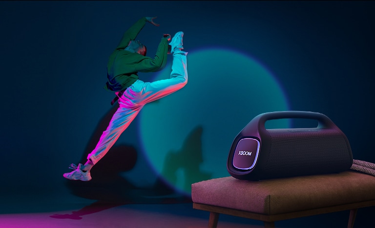 O LG XBOOM GO XG9 está colocado em uma cadeira. Ele projeta uma ilumina??o cênica na parede, e uma pessoa dan?a na frente dela.