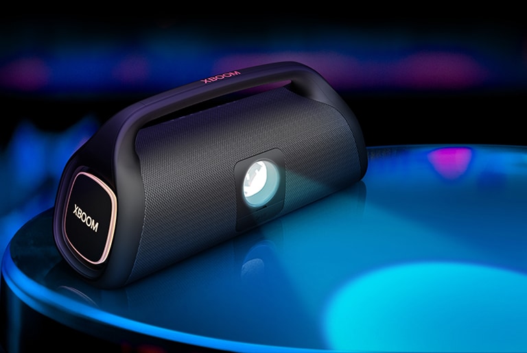 O LG XBOOM Go XG9 está colocado na superfície, mostrando sua lampada de ilumina??o cênica. A lampada projeta um uma luz azul e, na lateral, a ilumina??o laranja está acesa.