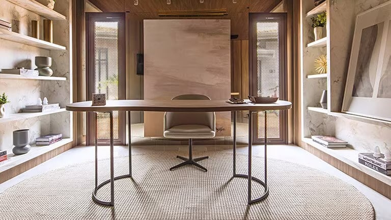 Home office com mesa oval de madeira e cadeira em tons de marrom no centro e nas laterais est?o as estantes de mármore repletas de objetos decorativos.