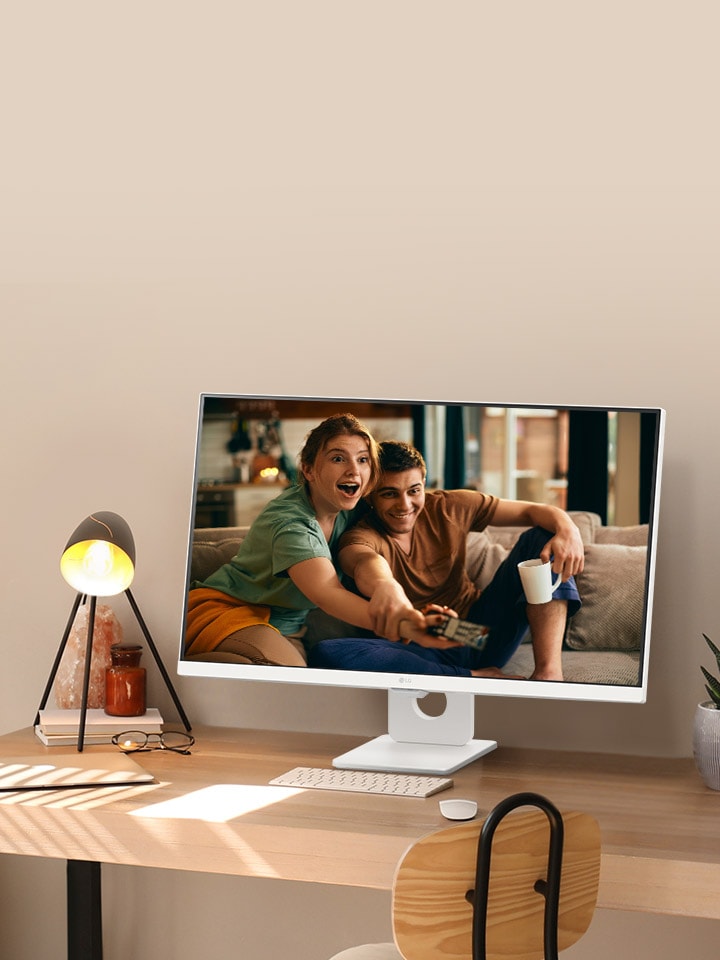 Na mesa perto da janela iluminada pelo sol, há um monitor inteligente exibindo um filme em sua tela. Ao lado do monitor, há uma lampada e um vaso de plantas. Na mesa, há um teclado e um mouse.