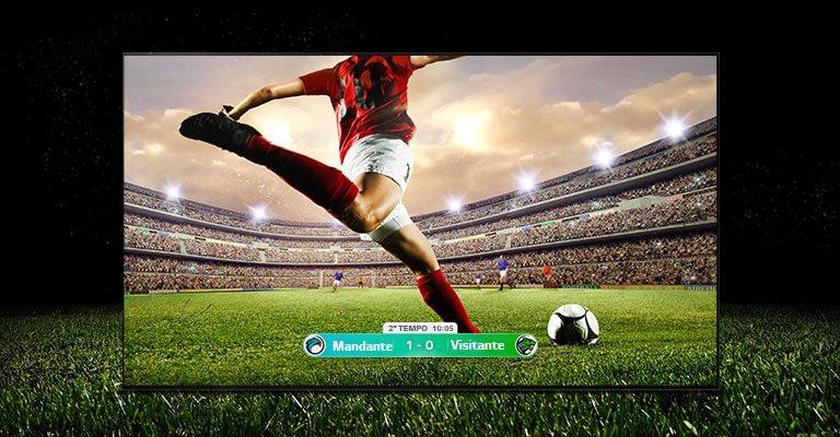 Uma imagem do display mostrando um jogo de futebol com um jogador de faixa vermelha prestes a chutar a bola para o outro lado do estádio. A pontua??o do jogo é visível na parte inferior da tela. A grama verde do campo se estende além da tela até o fundo preto.