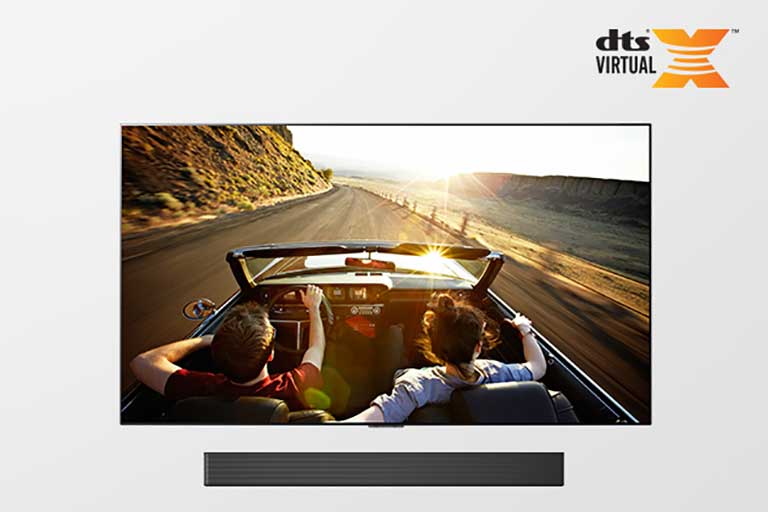 TV e Sound bar juntos em vista completa. A TV mostra um casal em um carro conversível numa estrada, rumo ao p?r do sol.