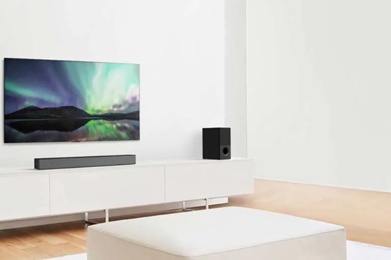 TV e Sound bar numa sala de estar branca com um sofá branco ao centro. Os alto-falantes est?o em ambas as extremidades do sofá.