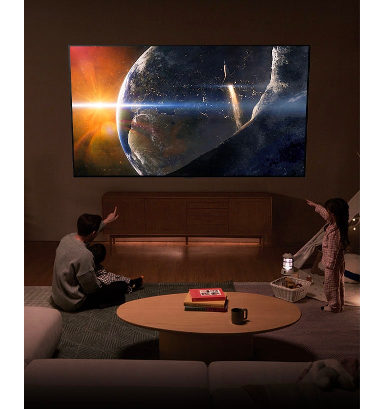 Uma família sentada no ch?o de uma sala de estar com pouca luz, ao lado de uma pequena mesa, olhando para cima para uma LG TV montada na parede, mostrando a Terra vista do espa?o.