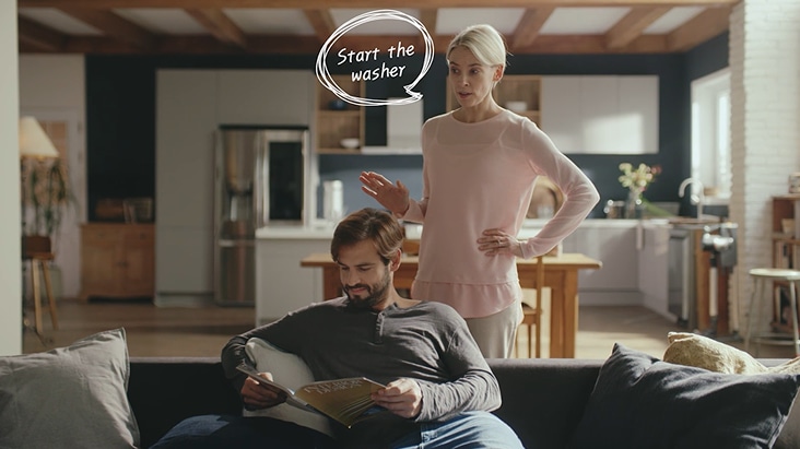 Em uma cozinha inteligente moderna, uma mulher usa um comando de voz para iniciar a lavadora com o LG ThinQ AI, enquanto um homem lê no sofá ao fundo.