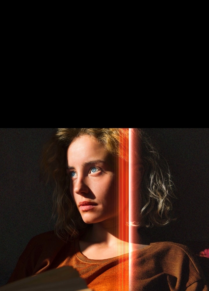 L’image d’une femme avec des yeux bleus per?ants et un chandail orange br?lé dans un coin sombre. Des lignes rouges démontrent des améliorations par IA qui couvrent une partie de son visage, illuminé dans les moindres détails, alors que le reste de l’image a l'air flou.