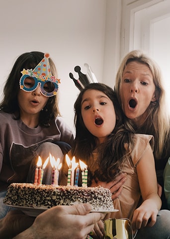 Bild von zwei erwachsenen Frauen und einem jungen M?dchen, das einen Geburtstagshut auf dem Kopf tr?gt und die Kerzen auf dem Kuchen ausbl?st.