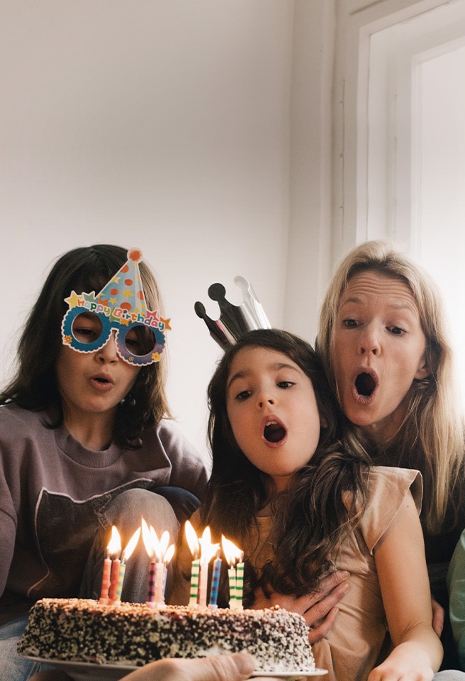 Image de deux femmes adultes et d’une jeune fille avec un chapeau d’anniversaire sur la tête et soufflant les bougies du gateau.