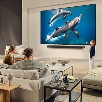 Una familia sentada en el sillón frente a la LG QNED TV montada en la pared encima de una LG Soundbar; una peque?a ni?a se?ala la pantalla que muestra dos delfines. El título habla de cómo las escenas son más emocionantes en una pantalla gigante.