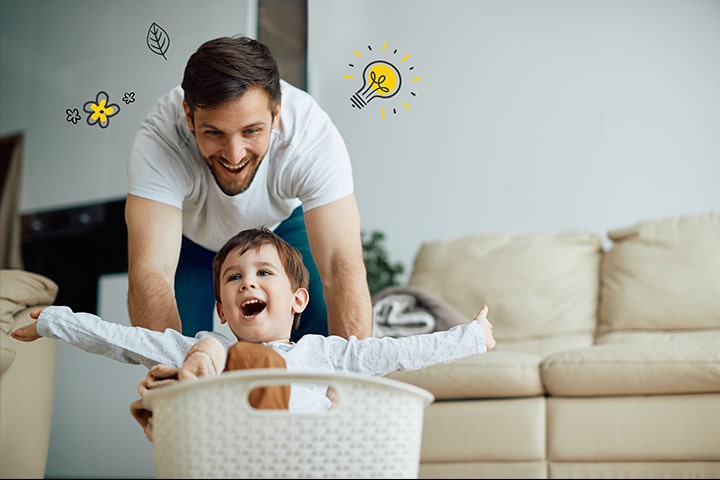 El padre empuja al ni?o en la cesta de la colada. A su alrededor hay una bombilla que simboliza el ahorro de energía, y una hoja y una flor que simbolizan la naturaleza.