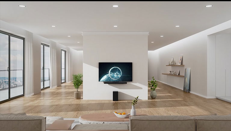 Soundbar a?televizor jsou zavě?eny na bílém sloupu uprost?ed obyvacího pokoje. Na d?evěné podlaze p?ímo pod soundbarem stojí ?erny subwoofer. Na levé straně jsou okna od podlahy a? ke stropu se skvělym vyhledem a?bílymi závěsy. Na pravé straně se nacházejí d?evěné police s?dekoracemi a?na podlaze stojí obraz.