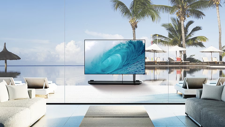 Televize LG SIGNATURE OLED TV W zobrazuje chladnou vlnu na obrazovce, zatímco je polo?ena v obyvacím pokoji s modrym vyhledem na oceán za oknem.