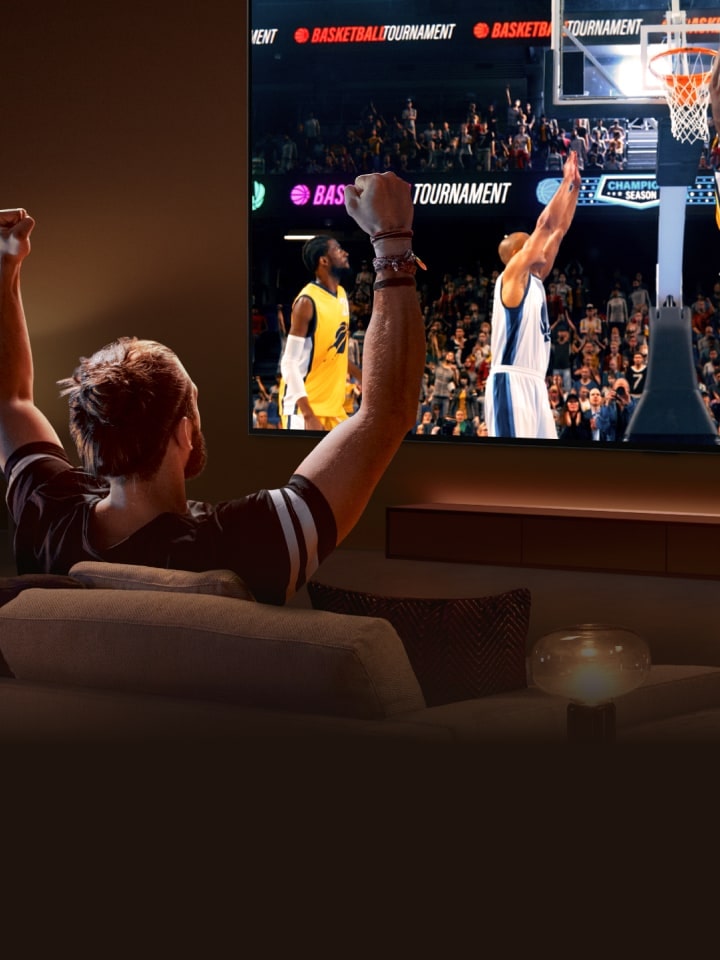 Un hombre sentado en un sofá frente a un televisor LG de gran tama?o que muestra un partido de baloncesto, con los brazos en alto como si estuviera animando.
