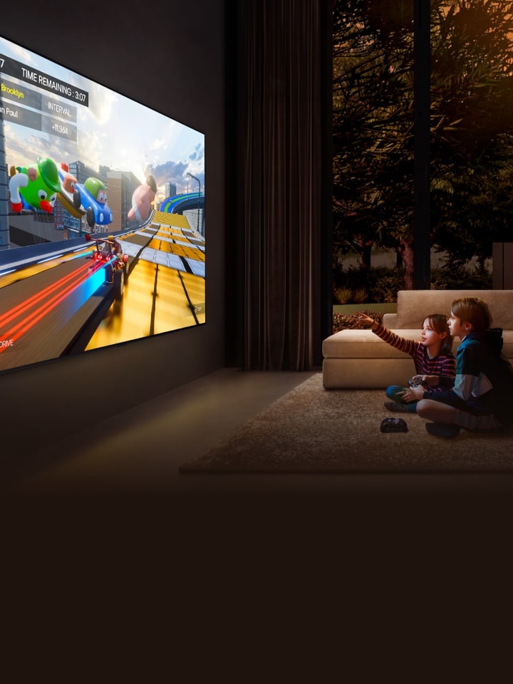 Vista en ángulo lateral de un televisor LG ultra grande montado en la pared de un acogedor salón que muestra un colorido juego, y dos ni?os sentados en el suelo frente al televisor sujetando los mandos del juego.