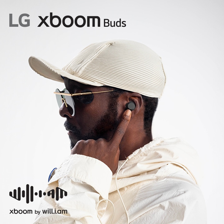 will.i.am in einem wei?en Outfit und mit Sonnenbrille zeigt nach rechts und deutet mit seinem linken Zeigefinger auf den Ohrh?rer in seinem Ohr.