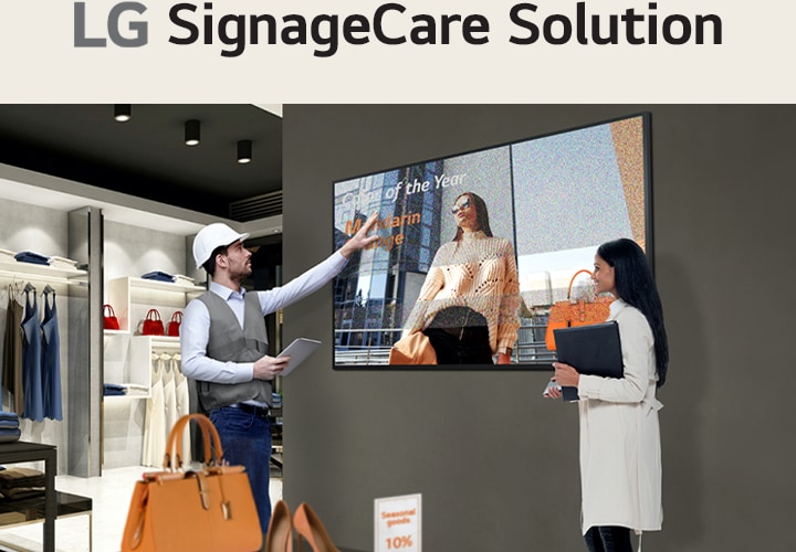 Ein Mitarbeiter eines Einzelhandelsgesch?fts in einer Weste und einem Schutzhelm zeigt auf ein digitales Display, das eine Frau mit dem Text ?Mitarbeiter des Jahres“ zeigt. Eine andere Person, die einen Ordner h?lt, beobachtet den Bildschirm in einem modernen Einzelhandelsbereich mit Regalen für Kleidung und Taschen.