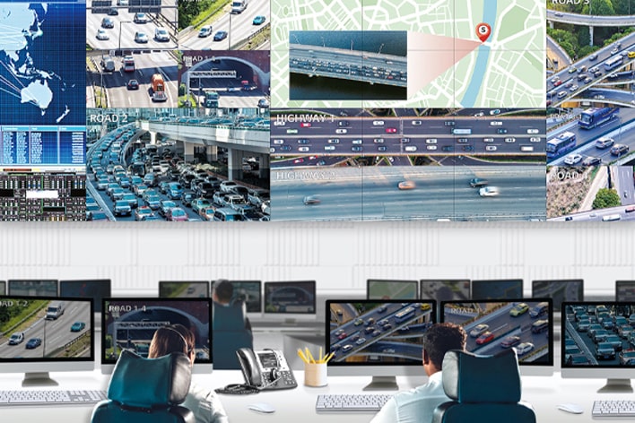 Ein Verkehrskontrollraum mit mehreren Monitoren, die Echtzeitaufnahmen von Autobahnen, Verkehrskarten und Analysen anzeigen. Zwei Bediener überwachen die Bildschirme, was fortschrittliche Verkehrsmanagement- und überwachungssysteme zeigt.