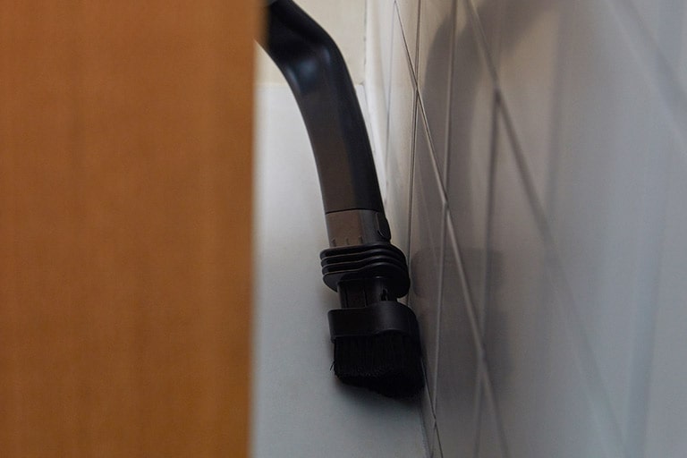 Der flexible Fugenaufsatz wird hinter einem M?belstück gezeigt, um Staub und Schmutz zwischen M?bel und Wand zu entfernen.