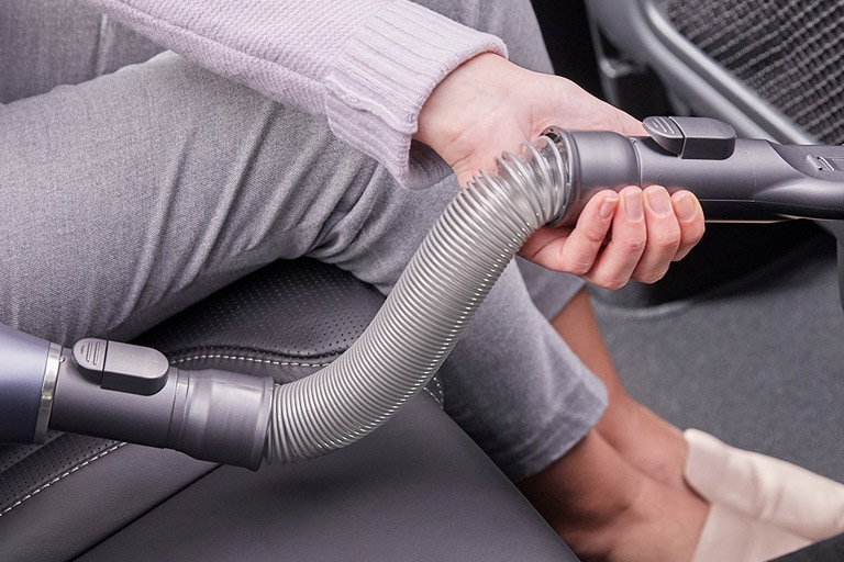 Man sieht die Beine, H?nde und Fü?e einer im Auto sitzenden Frau, w?hrend sie die flexible Schlauchverl?ngerung des Staubsaugers benutzt, um den Schmutz im Auto aufzusaugen.