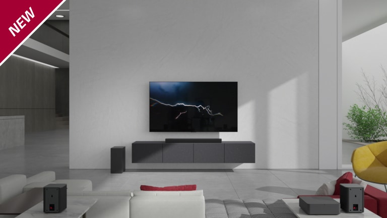 Eine Sound Bar steht in einem Wohnzimmer auf einem grauen TV-Regal unter einem wandmontierten Fernseher. Ein kabelloser Subwoofer steht links davon auf dem Boden und die Sonne scheint von rechts in den Raum. Ein langes Sofa in Rot und Wei? steht gegenüber dem Fernsehger?t und der Sound Bar. In der linken oberen Ecke steht ?NEU“.