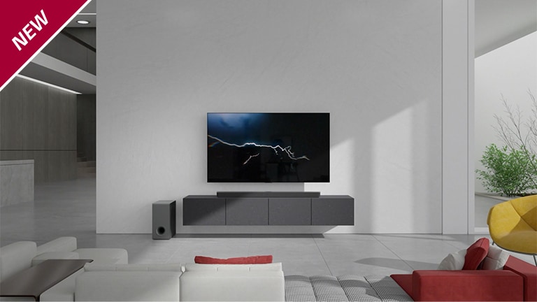 Eine Sound Bar steht in einem Wohnzimmer auf einem grauen TV-Regal unter einem wandmontierten Fernseher. Ein kabelloser Subwoofer steht links davon auf dem Boden und die Sonne scheint von rechts in den Raum. Ein langes Sofa in Rot und Wei? steht gegenüber dem Fernsehger?t und der Sound Bar. In der linken oberen Ecke steht ?NEU“.