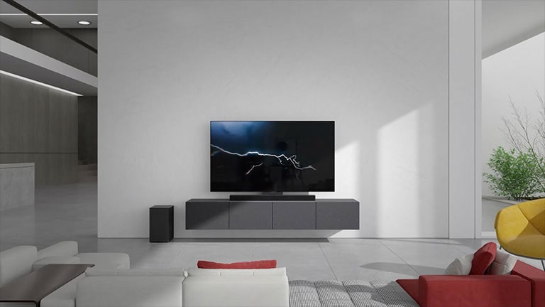 Die Soundbar steht im Wohnzimmer auf dem grauen Schrank mit einem Fernsehger?t. Ein schwarzer kabelloser Subwoofer steht auf dem Boden auf der linken Seite und die Sonne scheint von rechts ins Bild. Ein langes Sofa in Rot und Wei? steht gegenüber dem Fernsehger?t und der Soundbar.