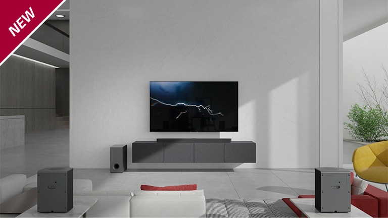 Eine Sound Bar steht in einem Wohnzimmer auf einem grauen TV-Regal unter einem wandmontierten Fernseher. Ein kabelloser Subwoofer steht links davon auf dem Boden und die Sonne scheint von rechts in den Raum. Ein langes Sofa in Rot und Wei? steht gegenüber dem Fernsehger?t und der Sound Bar. In der linken oberen Ecke steht ?NEU“.