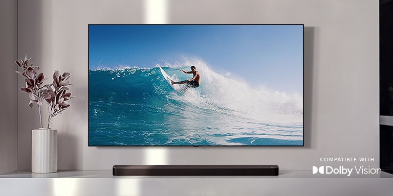 An der Wand befestigter TV. Der TV zeigt einen Mann, der auf einer gro?en Welle surft. Die LG Soundbar steht direkt unter dem TV auf einem wei?en Regal. Direkt neben der Soundbar befindet sich eine Vase mit einer Blume (Video abspielen).
