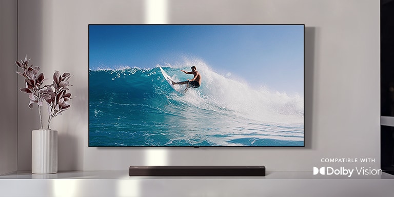 An der Wand befestigter TV. Der TV zeigt einen Mann, der auf einer gro?en Welle surft. Die LG Soundbar steht direkt unter dem TV auf einem wei?en Regal. Direkt neben der Soundbar befindet sich eine Vase mit einer Blume (Video abspielen).
