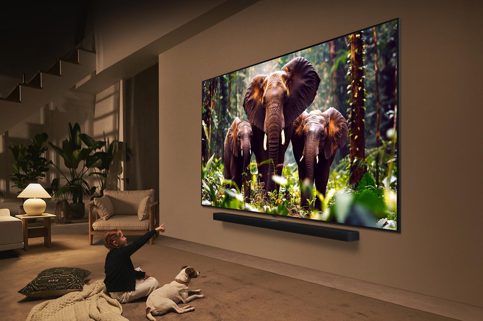 Ein M?dchen und ein Hund sitzen vor einem LG QNED-Fernseher, der an einer Wand montiert ist und drei Elefanten zeigt, die über einer LG Soundbar nach au?en laufen. Der Titel spricht davon, dass jede Aktion auf einem gro?en Bildschirm noch spannender ist.