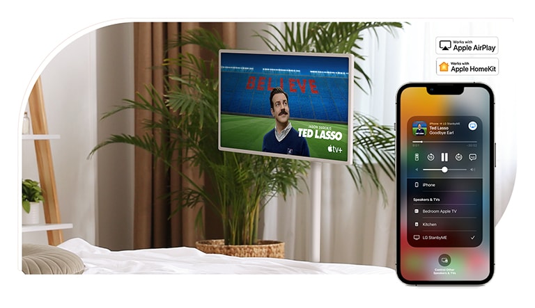 Ein Fernseher steht in einem gemütlichen Schlafzimmer und auf dem Bildschirm l?uft eine TV-Show - TED LASSO. Es gibt ein mobiles Ger?t auf dem gleichen Bild, das AirPlay UI in seinem Bildschirm zeigt. In der rechten oberen Ecke des Bildes sind das Apple AirPlay-Logo und das Apple HomeKit-Logo zu sehen.