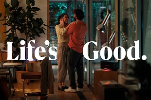 Ein l?chelndes Paar steht in einem warm beleuchteten, modernen Wohnzimmer mit bodentiefen Fenstern und Zimmerpflanzen. Gro?formatiger wei?er Text 'Life’s Good.' ist eingeblendet.