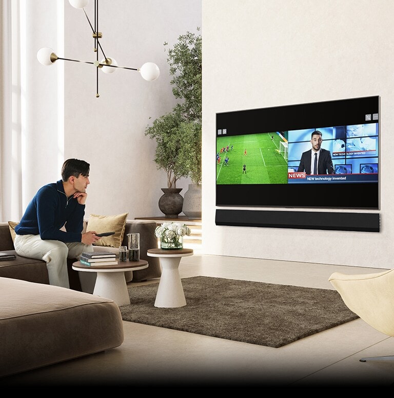 Ein Mann sitzt in einem gro?en, luxuri?s eingerichteten Wohnzimmer auf dem Sofa und sieht fern. Der Bildschirm ist zweigeteilt: Auf der linken H?lfte l?uft ein Fu?ballspiel, die rechte H?lfte zeigt eine Nachrichtensendung an.