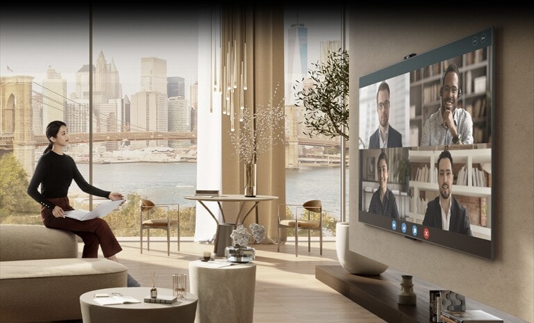 Eine Frau sitzt in einem gro?en, elegant eingerichteten Wohnzimmer auf der Couch. Sie hat ein ge?ffnetes Notebook auf dem Scho? und blickt als Teilnehmerin an einer Videokonferenz auf einen gro?en Fernseher an der gegenüberliegenden Wand.