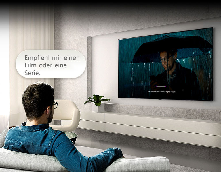 Ein Mann sitzt auf einem Sofa und blickt auf einen Fernseher. über seinem Kopf befindet sich eine Sprachblase mit den Worten ?Empfiehl mir einen Film oder eine Serie.“ Die Szene auf dem Bildschirm zeigt einen Mann mit Regenschirm, au?erdem ist die Benutzeroberfl?che der Spracherkennung zu sehen.