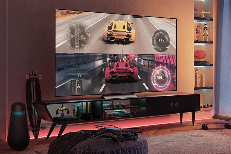 In einem Gaming-Raum mit r?tlicher Beleuchtung ist auf einem gro?en Fernseher ein Rennspiel mit gelben und roten Autos zu sehen. Links vom Fernseher steht ein spezieller Spiele-Lautsprecher, auch der Rest der Einrichtung ist ganz auf das Gaming abgestimmt.