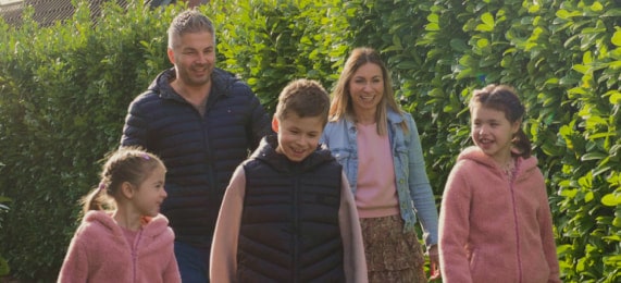 Eine fünfk?pfige Familie genie?t einen Spaziergang im Freien, l?chelt und tr?gt legere Kleidung, umgeben von grünen Hecken.