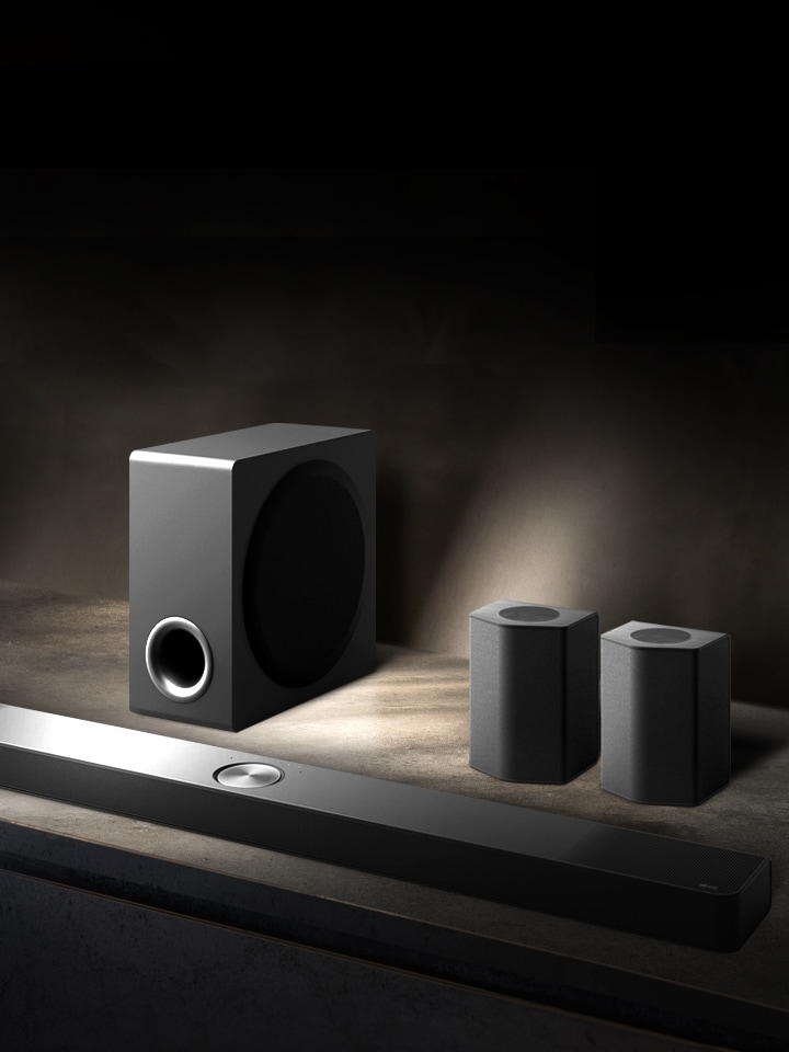 Visning oppefra af LG Soundbar, bagh?jttalere og bash?jttaler p? en brun tr?hylde i et sort rum, indhyllet i m?rke, hvor der kun er lys p? lydsystemet.	
